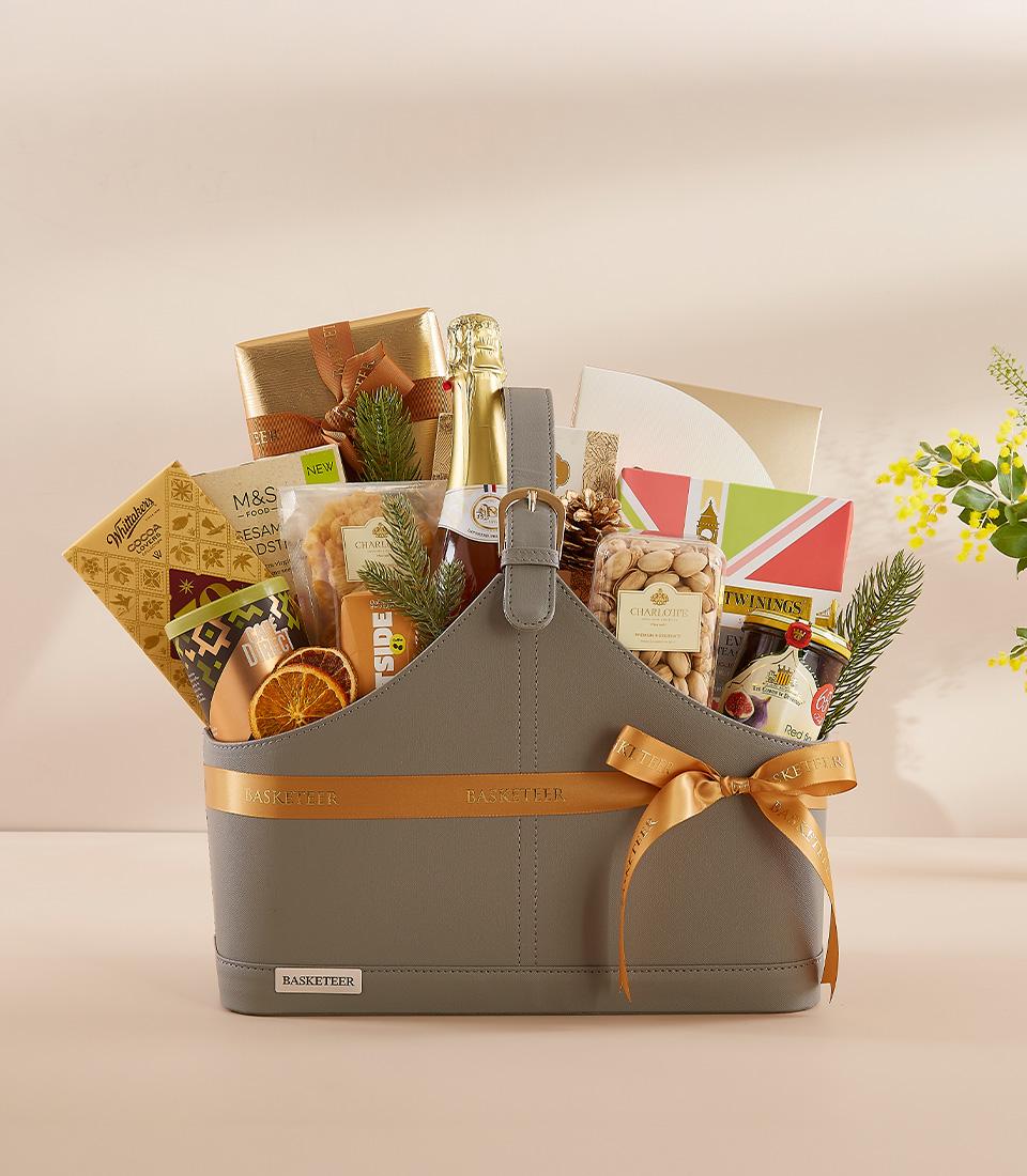 Grey faux leather gourmet gift basket with gold ribbon, sparkling beverage, chocolates, tea, and festive foliage for New
