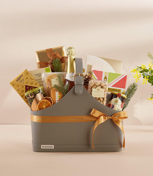 Grey faux leather gourmet gift basket with gold ribbon, sparkling beverage, chocolates, tea, and festive foliage for New