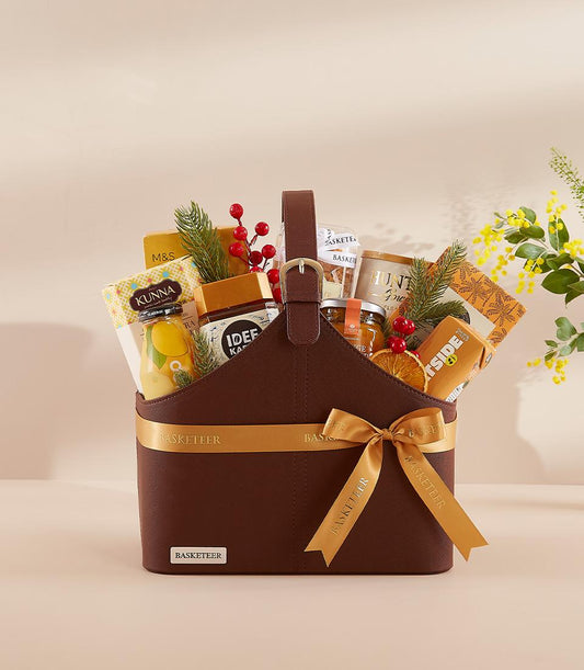 Dark brown luxury gift basket with gold buckle and ribbon, filled with gourmet jars, treats, and festive greenery