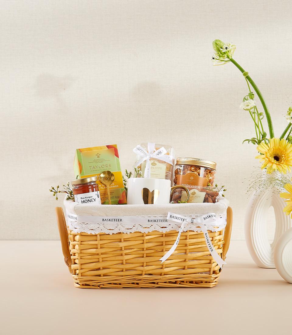 Luxury wicker gift basket with gourmet tea, honey, popcorn, cookies, gold-heart mug, and fresh flowers for New Year wellness