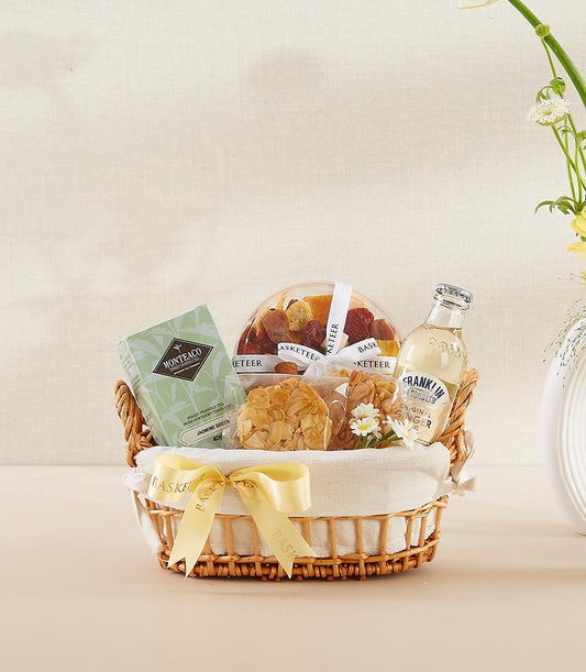 Woven wicker gift basket with jasmine green tea, dried fruits, ginger beverage, and almond florentine cookies, tied with
