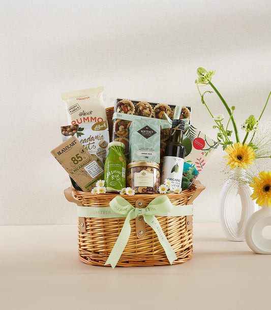 Woven wicker gourmet gift basket with macadamia nuts, chocolate, jasmine tea, olive oil, and pastries, tied with green ribbon