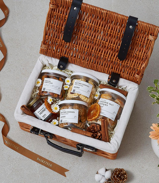 Light wicker picnic basket with butter cookies, caramel corn flakes, cashew cookies, and honey jar nestled in shredded paper