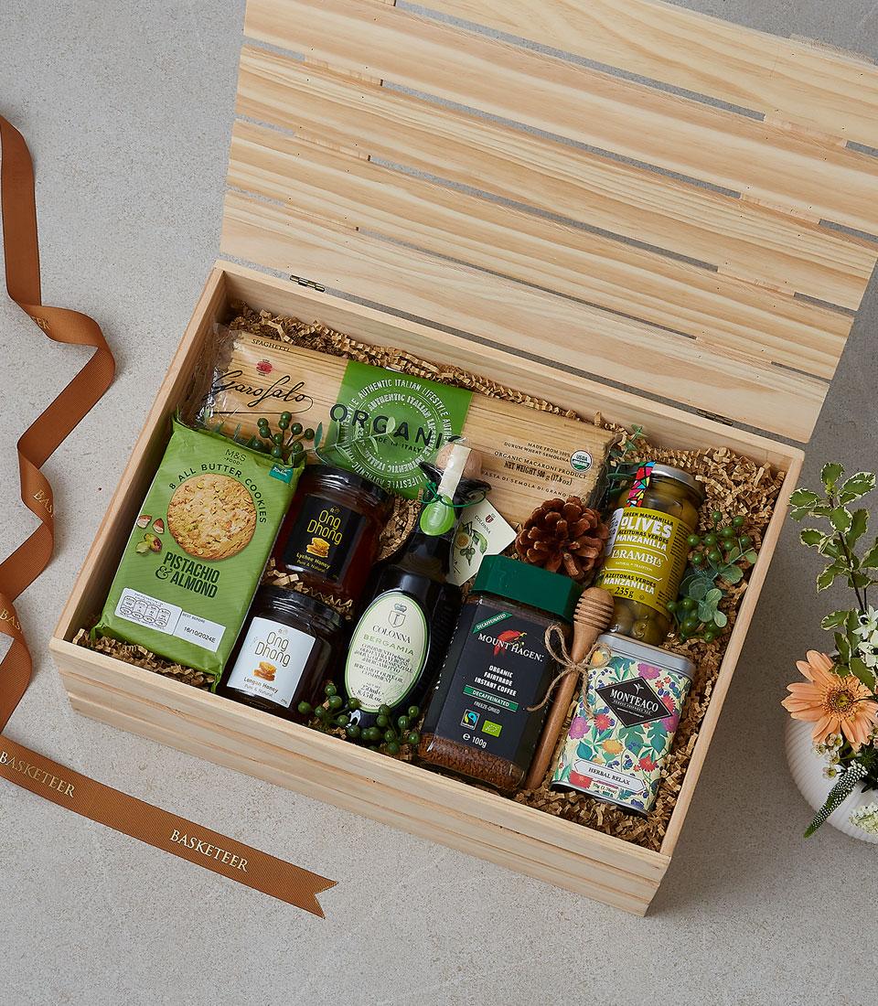 Open wooden gourmet gift box with artisan olive oil, honey jars, cookies, coffee, and tea on shredded paper