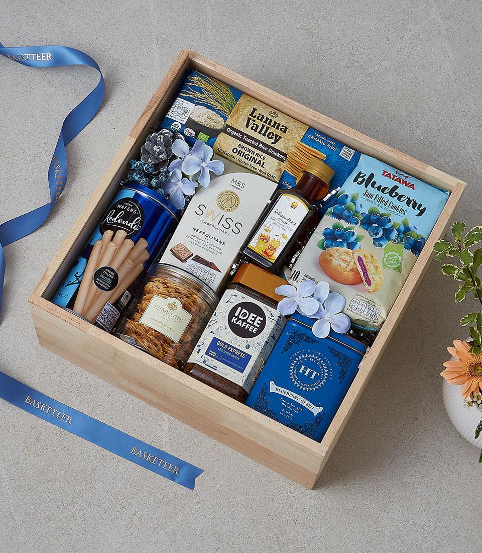 Wooden gift box filled with gourmet snacks, coffee, tea, chocolates, and jam cookies with blue ribbon