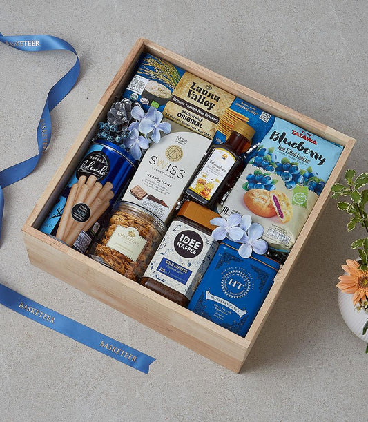 Wooden gift box filled with gourmet snacks, coffee, tea, chocolates, and jam cookies with blue ribbon