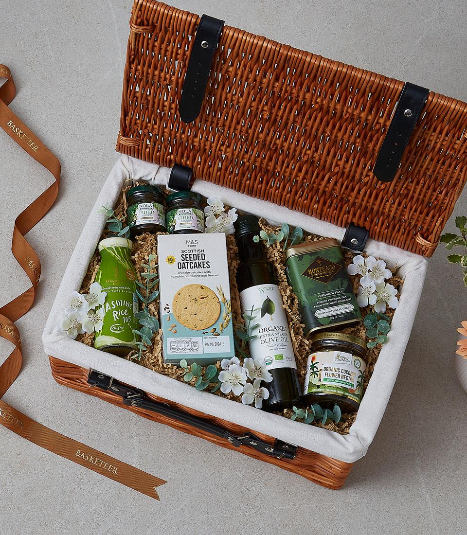 Woven wicker picnic basket filled with gourmet foods, artisan oils, and organic treats with white flowers and green accents