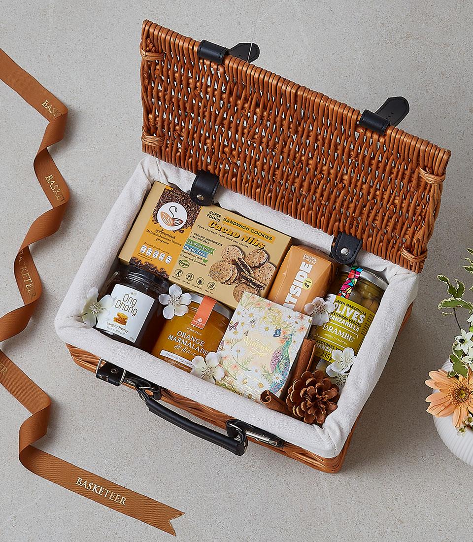 Light brown wicker picnic basket with gourmet treats, honey, marmalade, olives, and winter florals
