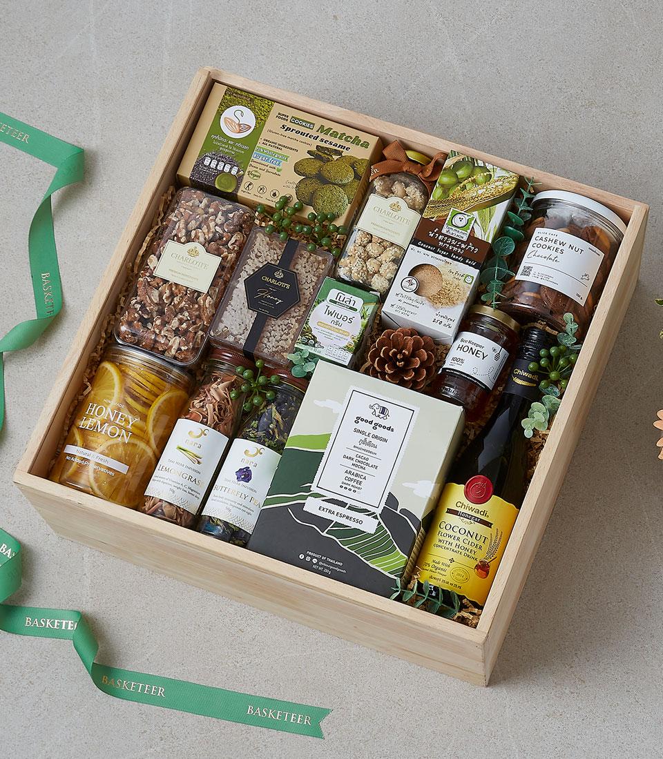 Rustic wooden gift box filled with gourmet snacks, honey, dried herbs, coffee, and matcha cookies tied with green ribbon