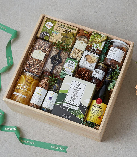 Rustic wooden gift box filled with gourmet snacks, honey, dried herbs, coffee, and matcha cookies tied with green ribbon