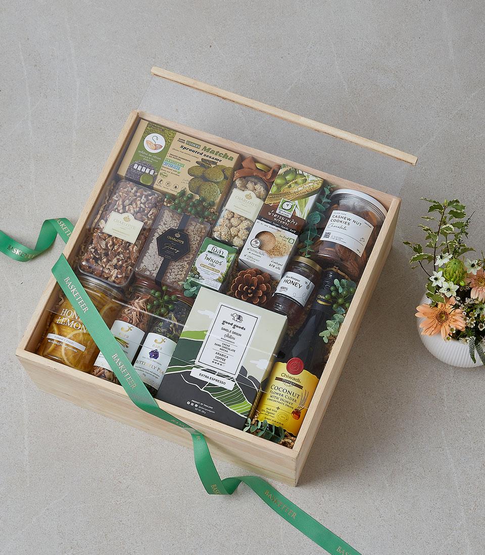 Light wooden gift box with green ribbon containing gourmet nuts, cookies, honey, dried fruit and coconut cider, adorned with
