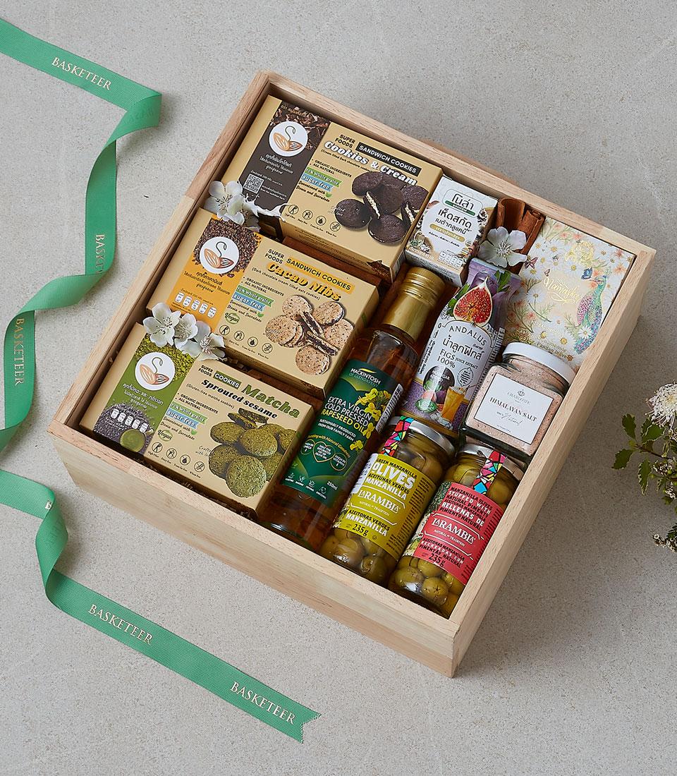 Wooden gift box filled with gourmet cookies, premium oils, olives, and artisan food items with white flowers and green