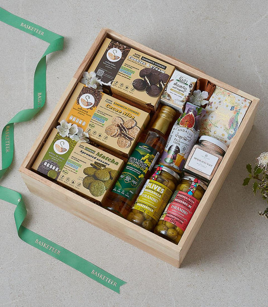 Wooden gift box filled with gourmet cookies, premium oils, olives, and artisan food items with white flowers and green