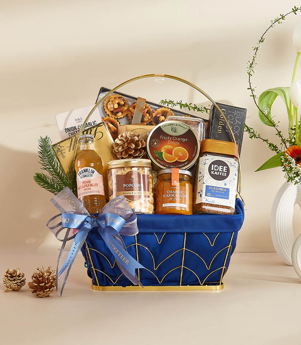 Elegant blue fabric gift basket with gold metal frame filled with gourmet treats, pink grapefruit soda, marmalade, and