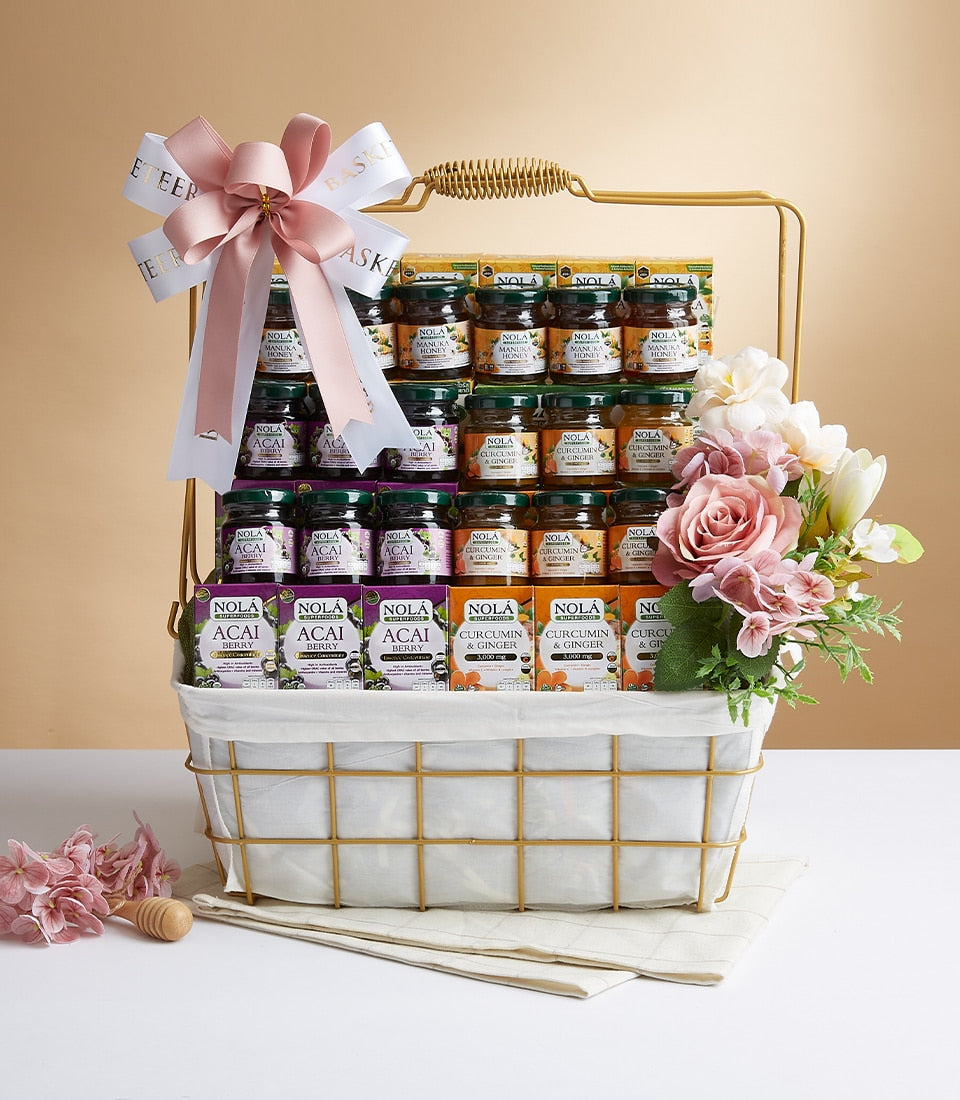 Gold wire gift basket with Nola wellness products, manuka honey jars, superfoods boxes, and fresh pink and white flowers