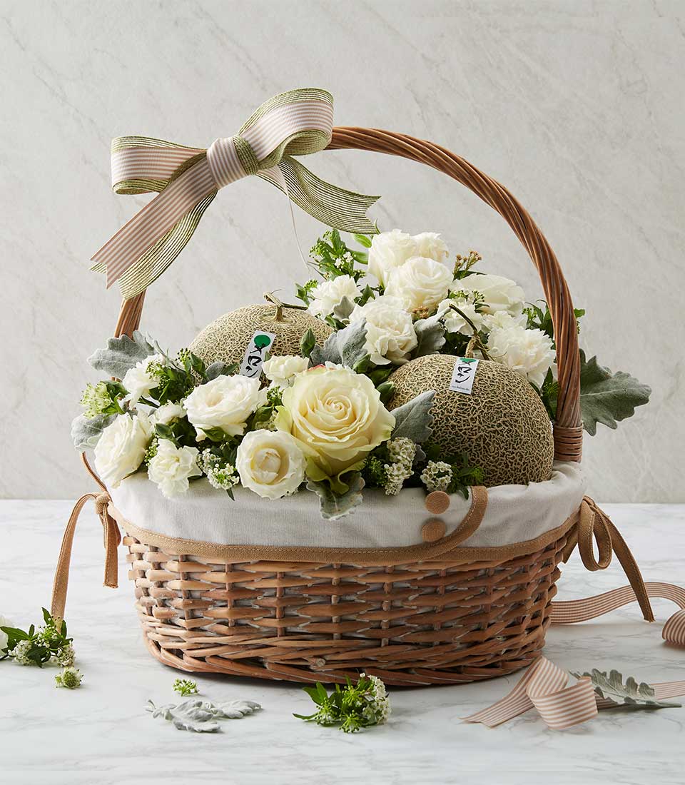 Light brown wicker gift basket with white flowers, fresh cantaloupes, and pale pink striped ribbon bow