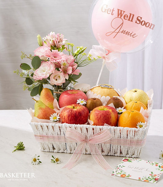 White woven fruit basket with fresh apples, pears, kiwis and citrus, pink-white flowers, and Get Well Soon balloon