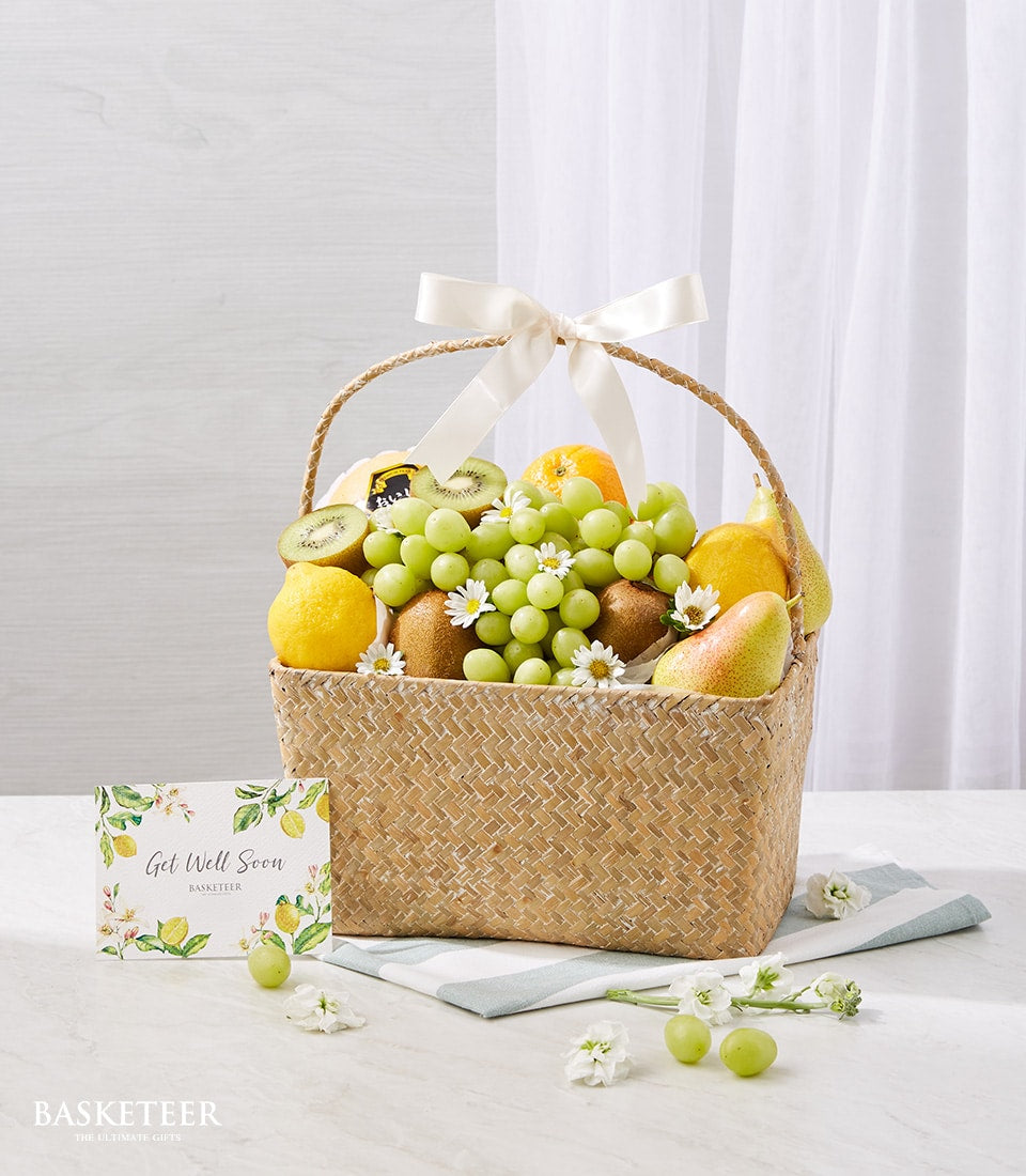 Woven wicker fruit basket with fresh grapes, kiwis, lemons, oranges, pears and white daisies, cream ribbon handle