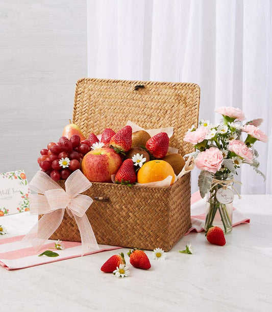 Woven straw fruit basket with fresh grapes, apples, strawberries and kiwis, paired with pink carnations and white daisies