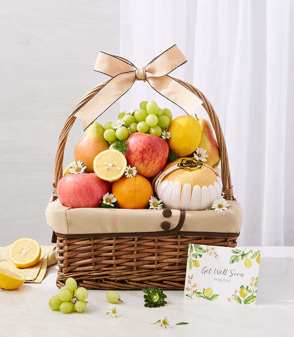 Woven brown wicker fruit basket with fresh grapes, apples, lemons, oranges, and pears, finished with peach ribbon bow