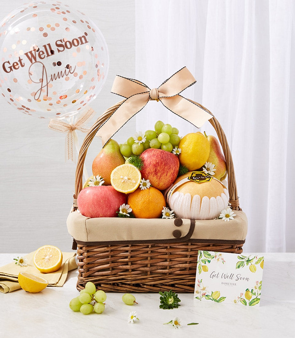 Wicker fruit basket with fresh apples, grapes, lemons, and oranges, beige ribbon bow, white daisies, Get Well Soon balloon