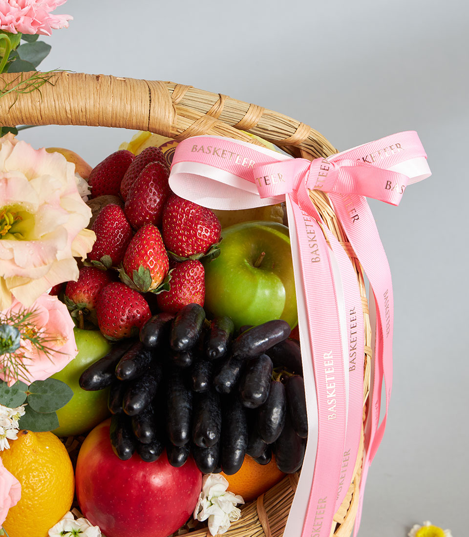 Pastel pink and cream floral arrangement with fresh strawberries, grapes, and citrus in woven wicker basket, perfect for