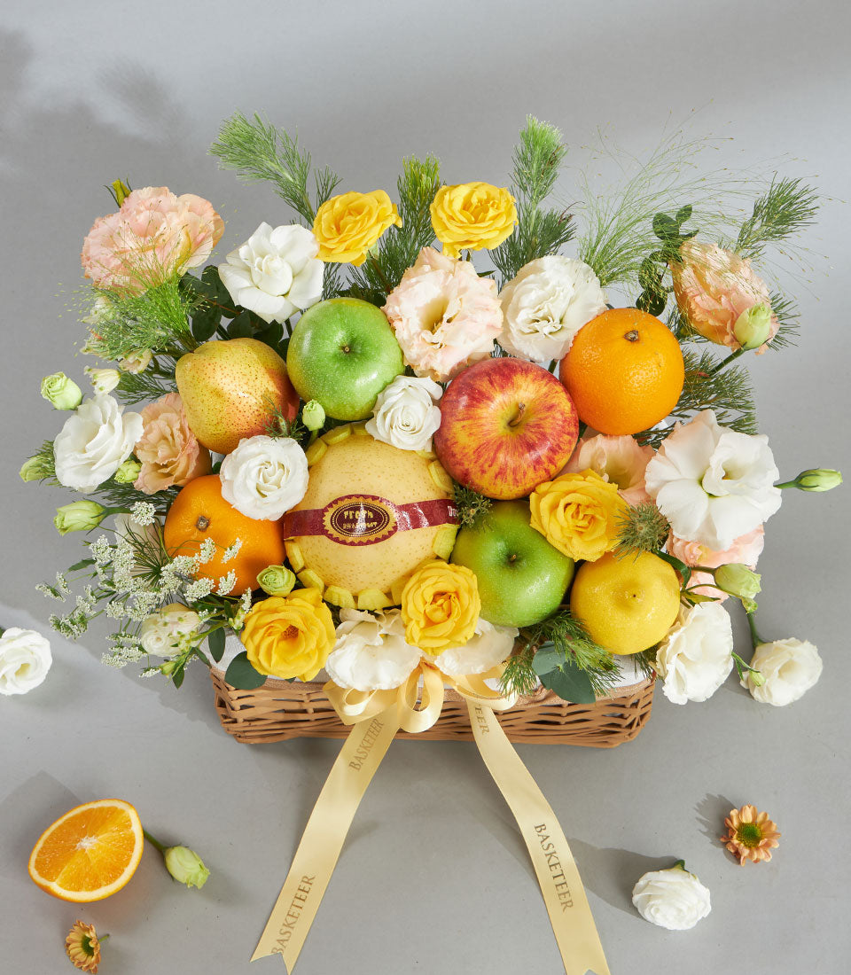 Light brown wicker basket filled with fresh yellow and red apples, pears, citrus, and mixed white and pale pink roses with