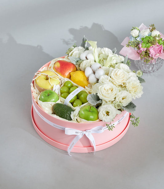 Pink round gift box with fresh fruit and white roses, calla lilies, and greenery arrangement for elegant gifting