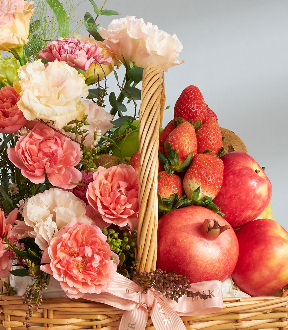 Basketeer luxury gift basket Bangkok, filled with fresh strawberries, apples, and pomegranates. A pretty peach ribbon accents the wicker hamper and floral arrangement.