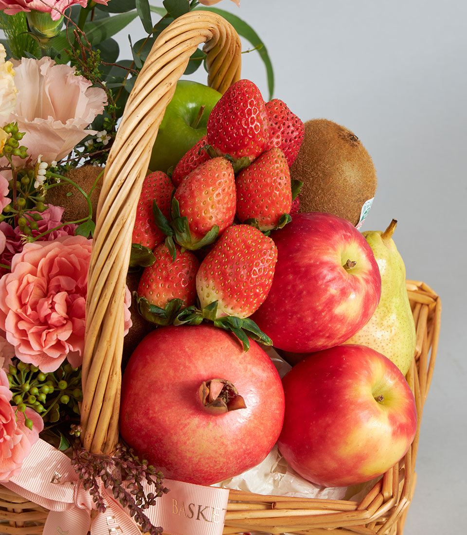 Basketeer premium fruit gift basket with apples, pomegranates, and strawberries, nestled amongst peach-toned florals in a wicker basket with light pink ribbon.