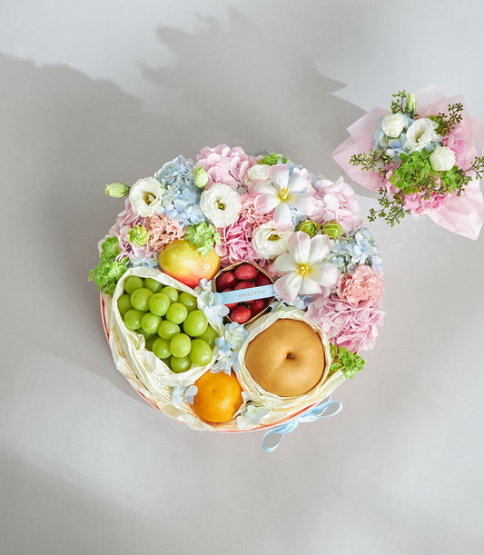 Soft pastel round gift box with fresh strawberries, pears, grapes, and orange surrounded by pink hydrangeas, blue eustoma,