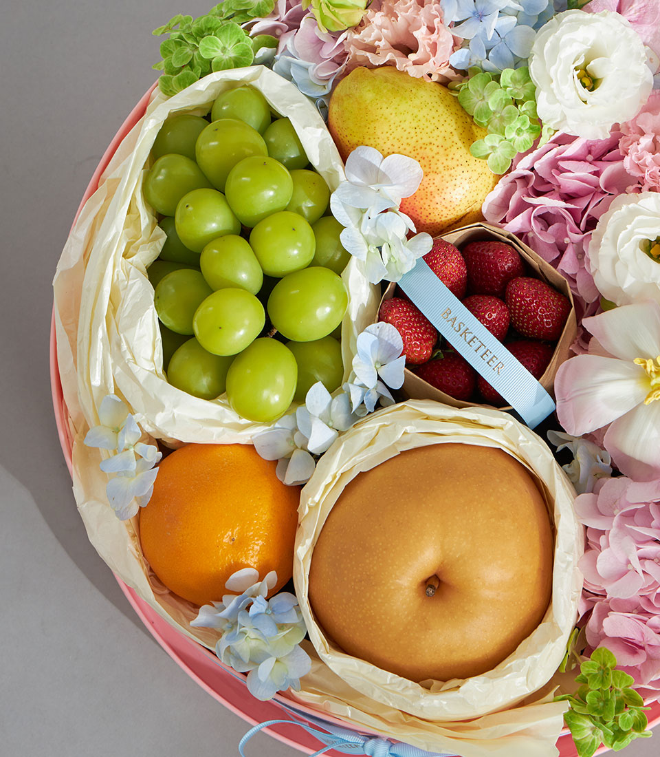 Pastel pink round gift box with fresh fruits, berries, and soft pink and blue flowers for birthday celebrations