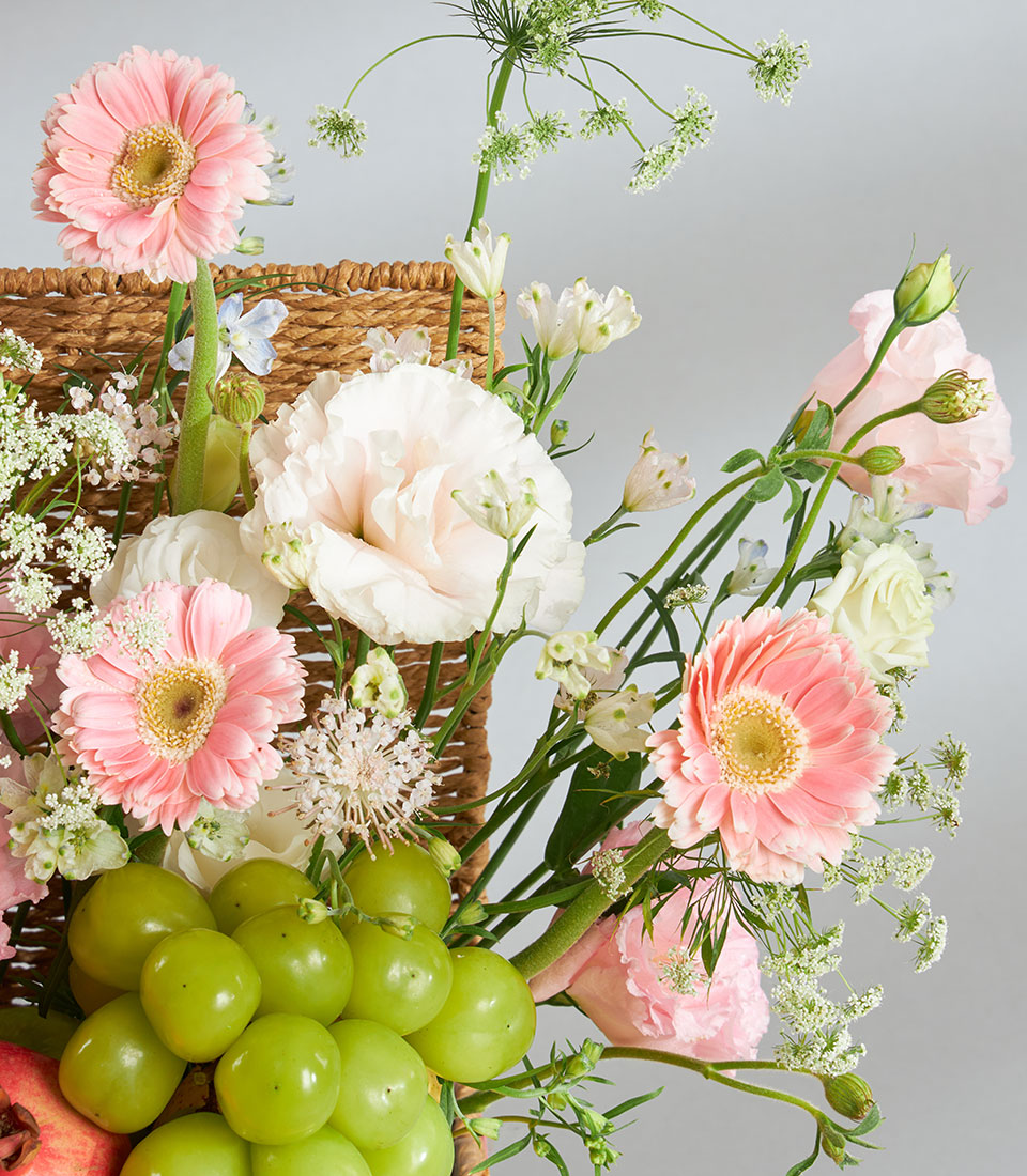 Premium pink pastel fruit and floral arrangement in woven basket with gerberas, lisianthus, and fresh grapes