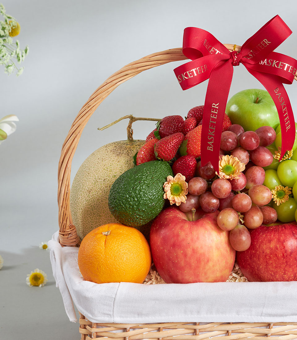 Premium mixed imported fruit basket with cantaloupe, apples, grapes, strawberries, and fresh florals in wicker