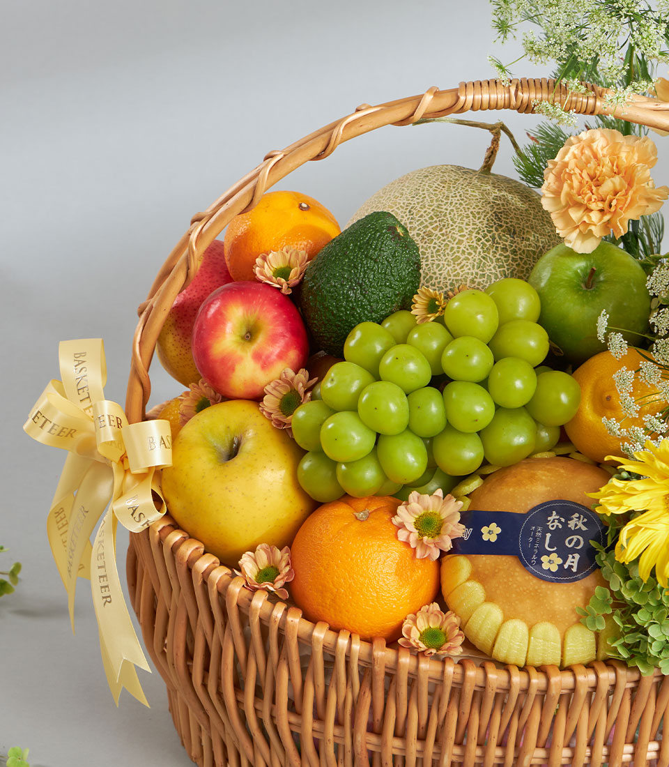 Wicker fruit basket with fresh apples, oranges, grapes, avocado and delicate yellow flowers, tied with yellow Basketeer