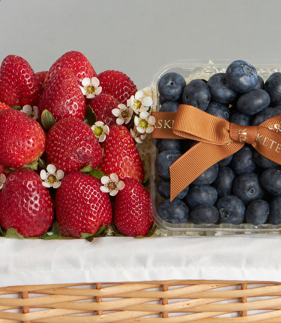 Fresh strawberries and blueberries in a wicker gift basket with white fabric lining and golden ribbon bow