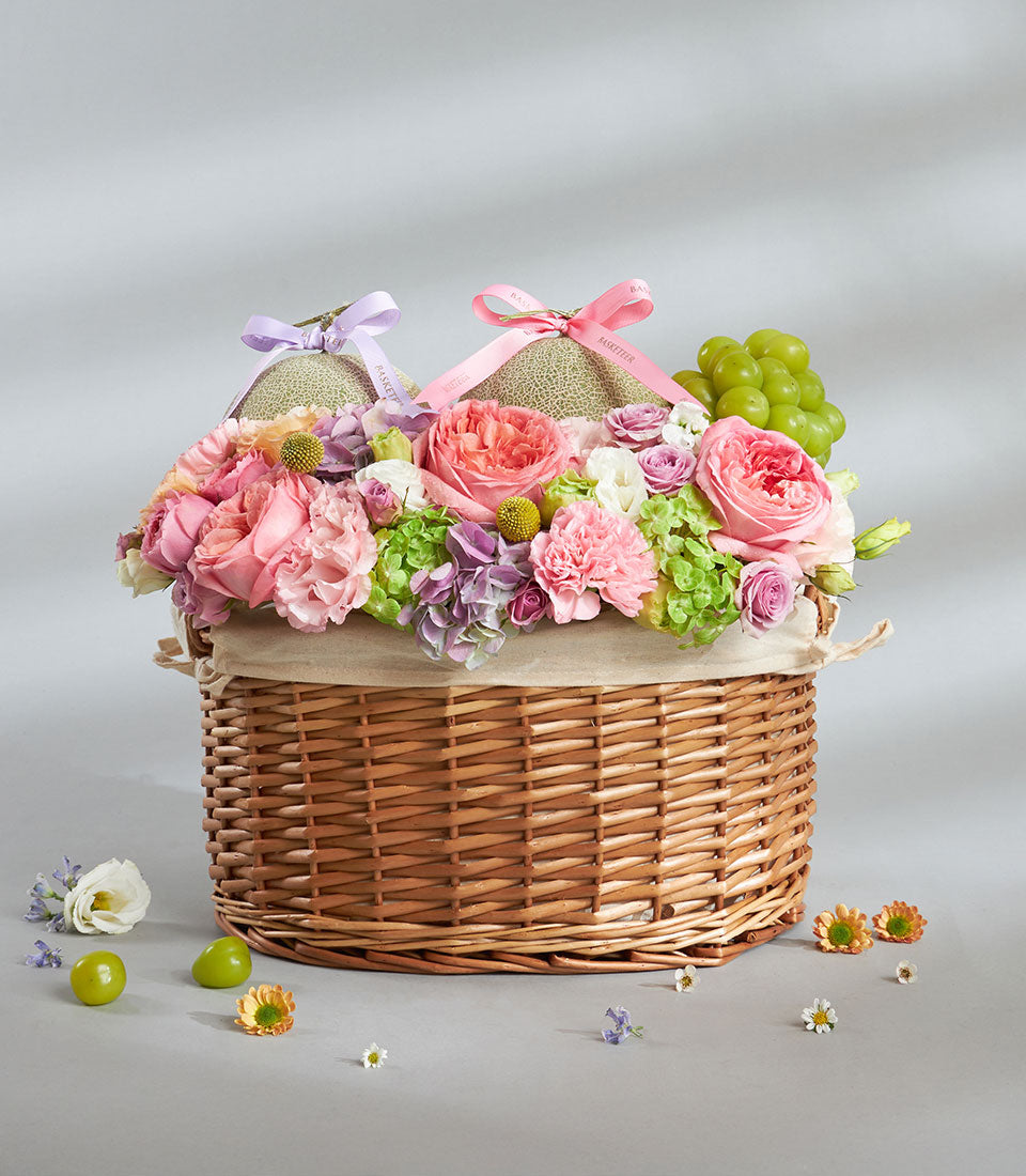 Elegant wicker gift basket with pastel melons, pink and peach roses, hydrangeas, and fresh grapes