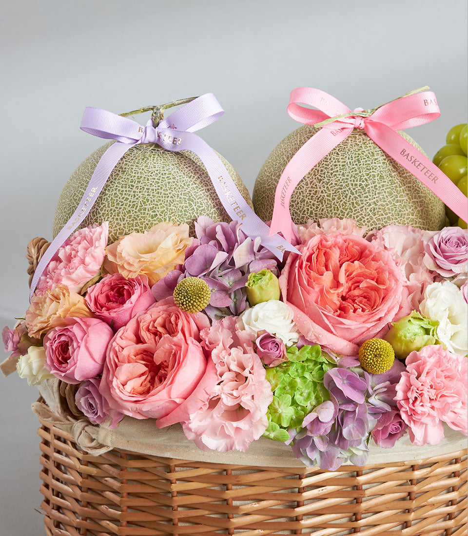 Pastel pink and purple melon duo gift basket with fresh roses, hydrangeas, and carnations in woven wicker