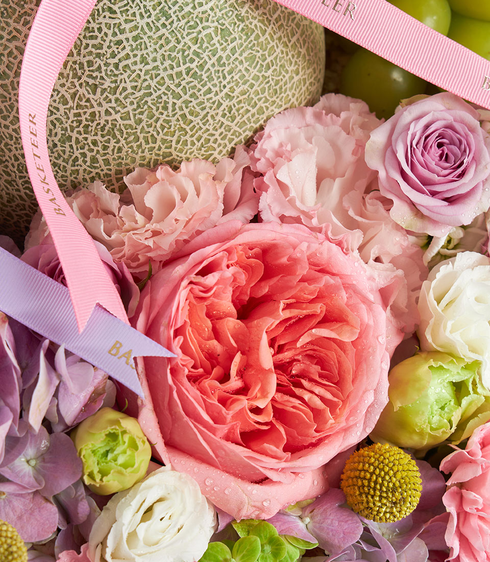 Elegant pastel gift basket with fresh melons, pink roses, hydrangeas, and green apples tied with silk ribbon