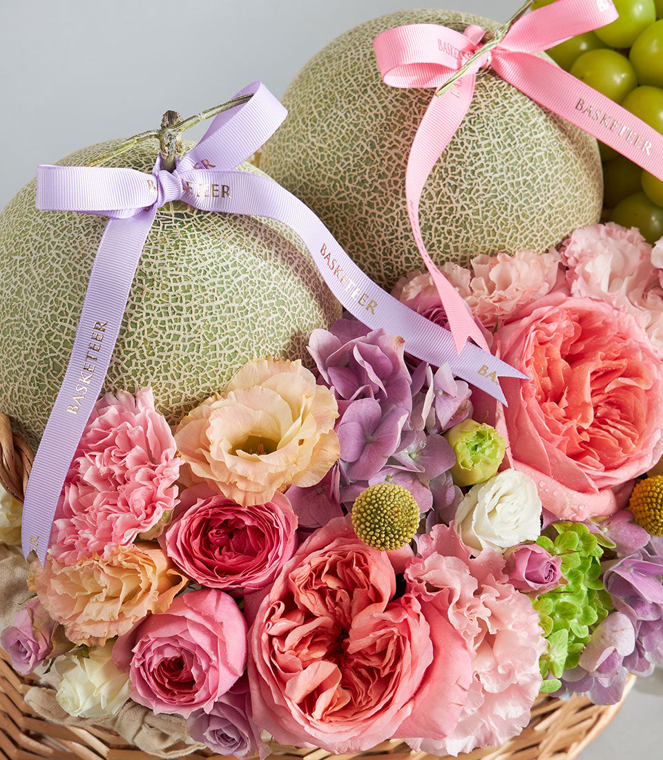 Luxury pastel gift basket with fresh cantaloupe melons, roses, hydrangeas, and seasonal fruits in woven basket
