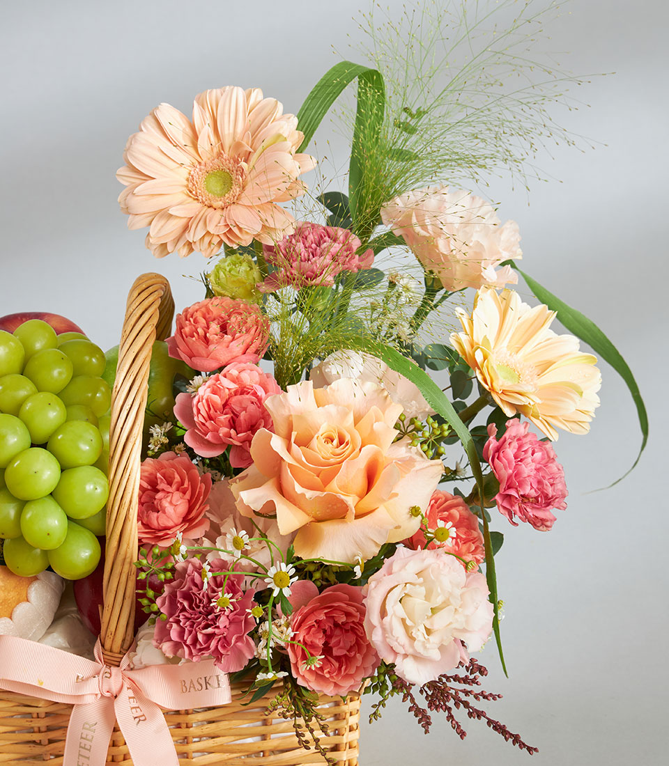 Peach and pink gerbera daisies, roses, and fresh fruit in a wicker birthday gift basket with ribbon bow