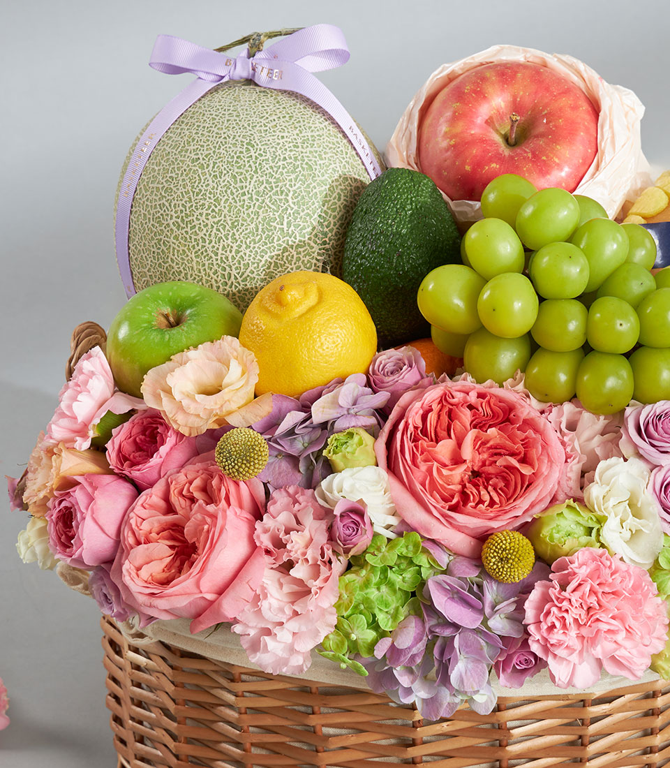 Woven wicker fruit basket with green cantaloupe, apples, grapes, and avocado arranged with pink, peach, and purple roses and