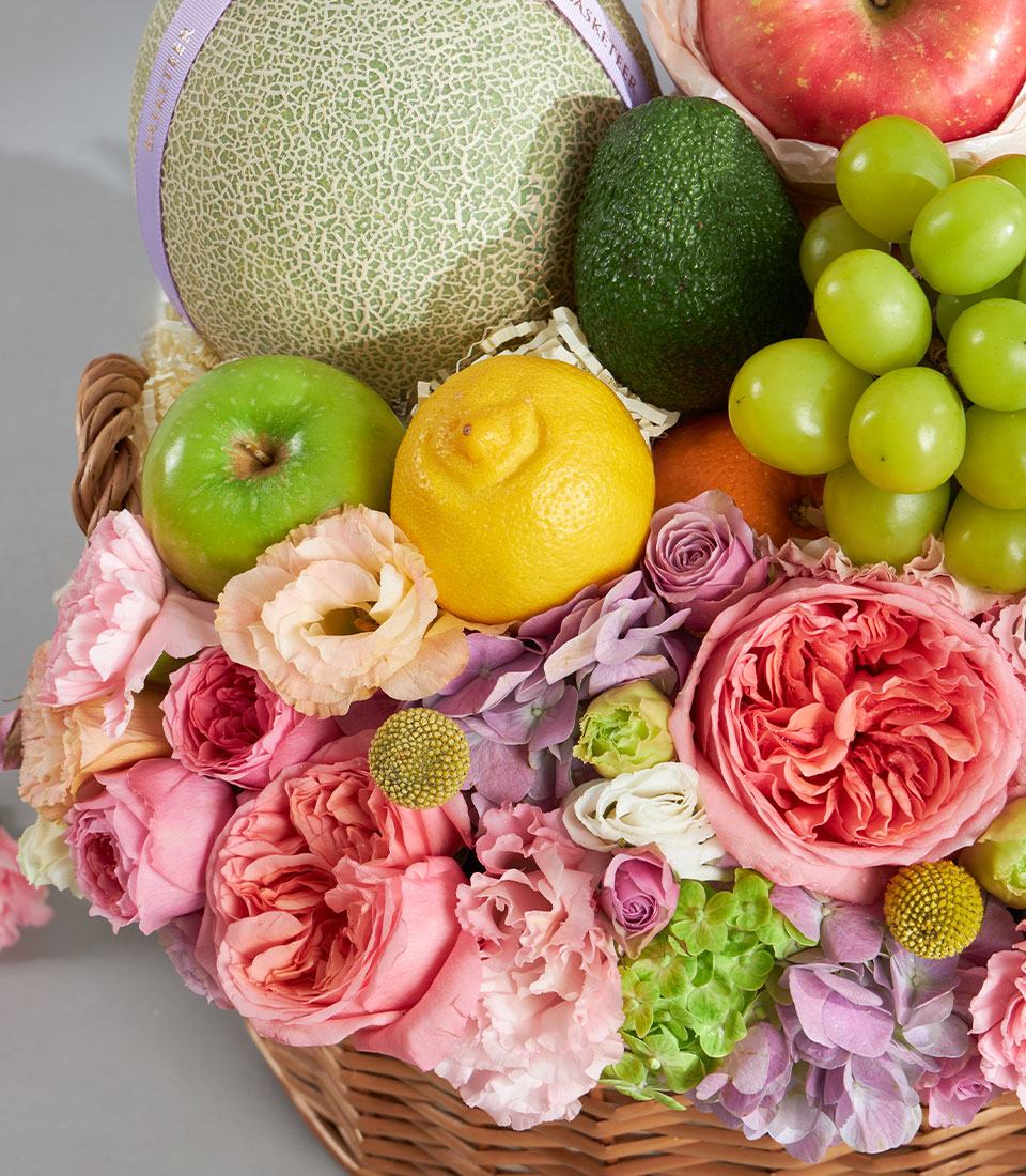 Pastel fruit and floral gift basket with cantaloupe, apples, grapes, and fresh roses in pink, peach, and purple