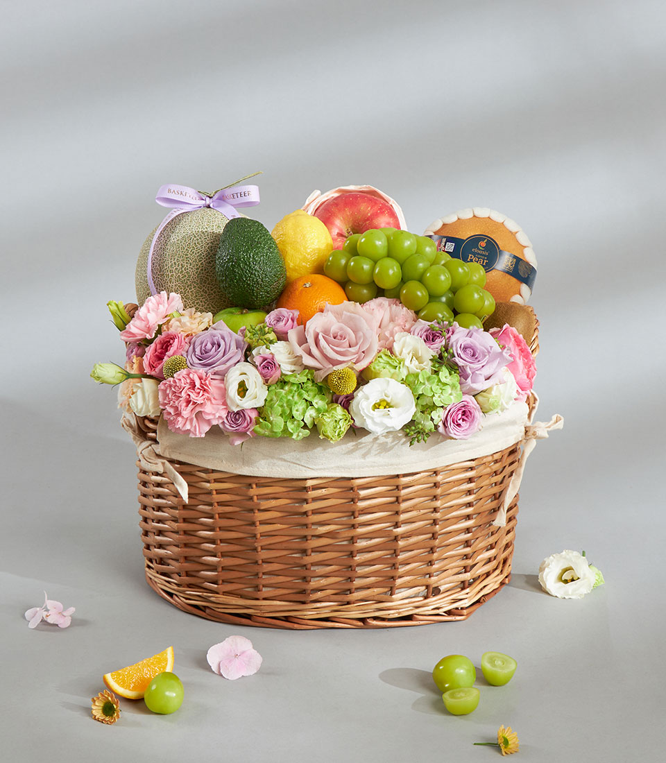 Pastel fruit and floral birthday basket with melon, grapes, and fresh pink and purple flowers in woven wicker