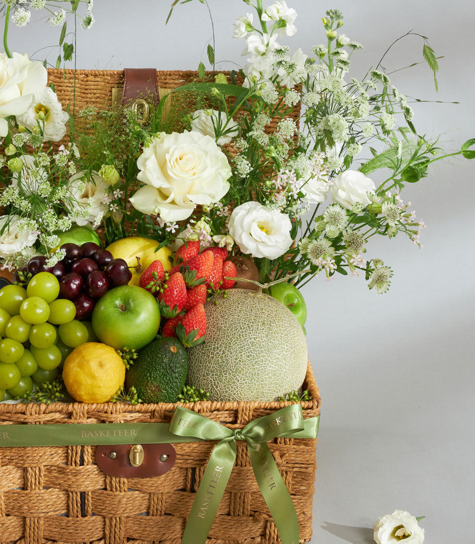Woven wicker fruit and flower basket with white roses, fresh produce, and green satin ribbon