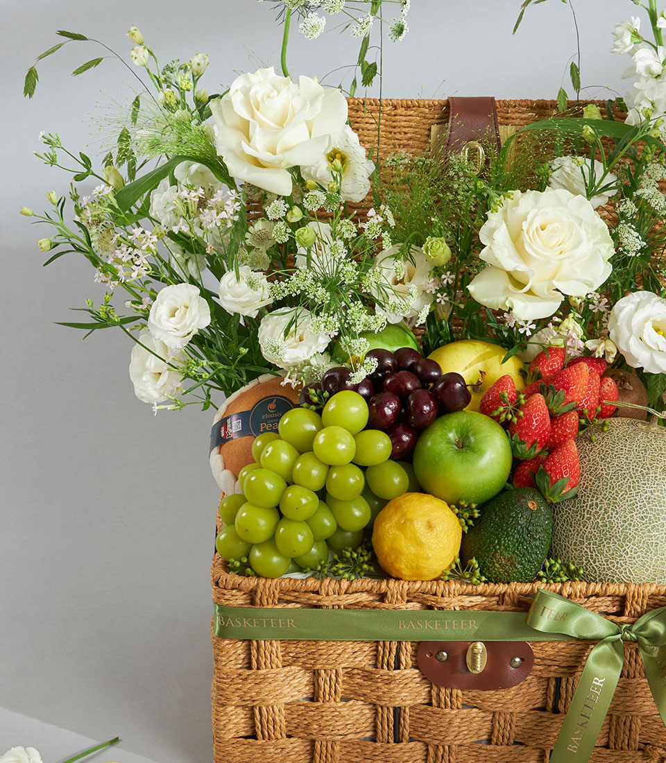 Woven fruit and flower basket with white roses, fresh berries, tropical fruits, and green foliage, tied with signature green