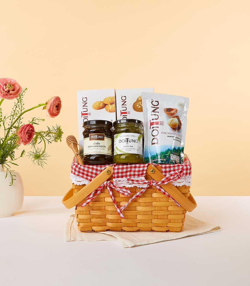 Woven picnic basket with gingham lining filled with Doi Tung artisan spreads, cookies, and macadamia nuts, styled with pink