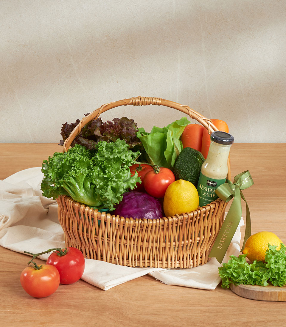 Wicker basket filled with fresh vegetables, lettuce, tomatoes, cabbage, carrots, avocado, lemon, and creamy dressing
