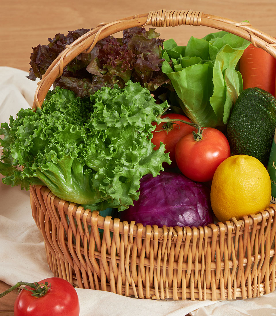Wicker basket overflowing with fresh vegetables, lettuce, tomatoes, purple cabbage, and citrus for health-conscious gifting