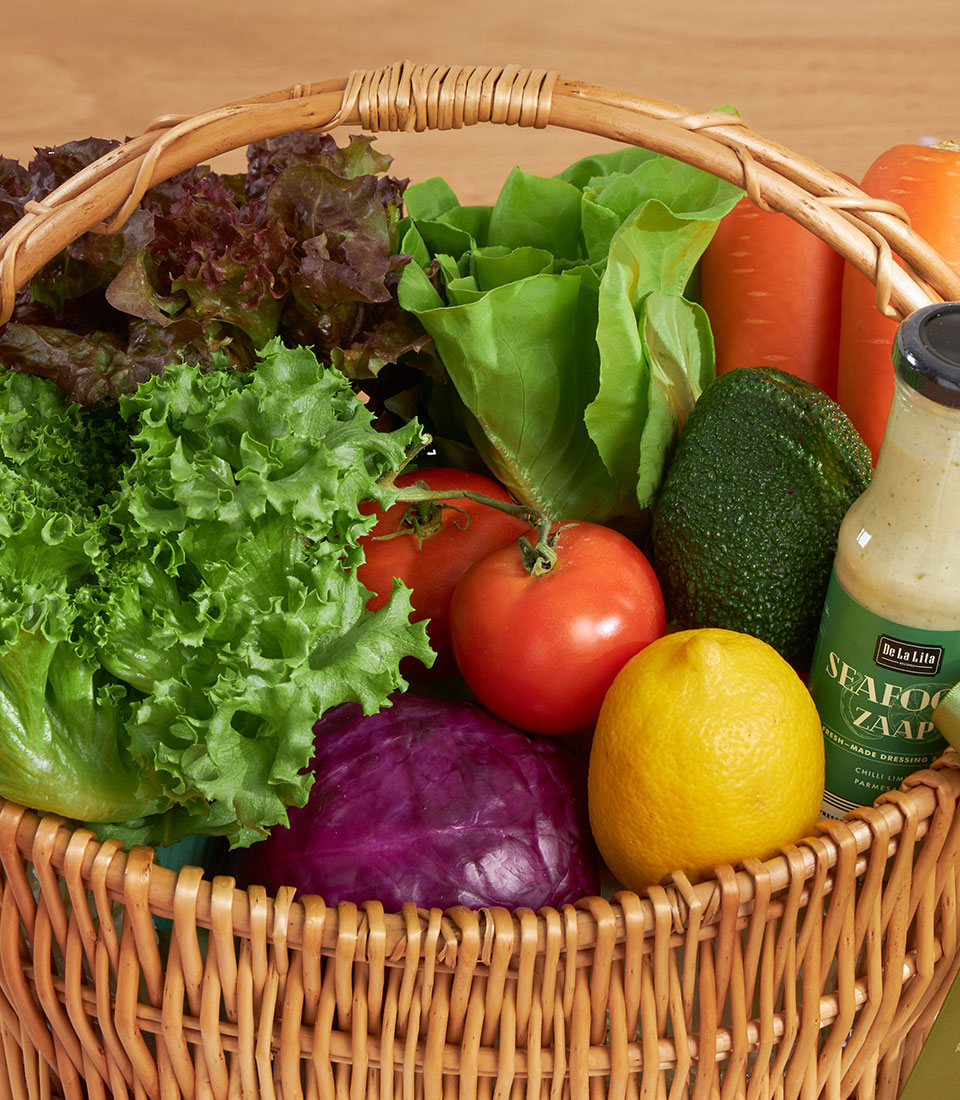 Wicker basket filled with fresh vegetables, leafy greens, tomatoes, carrots, avocado, cabbage and homemade dressing